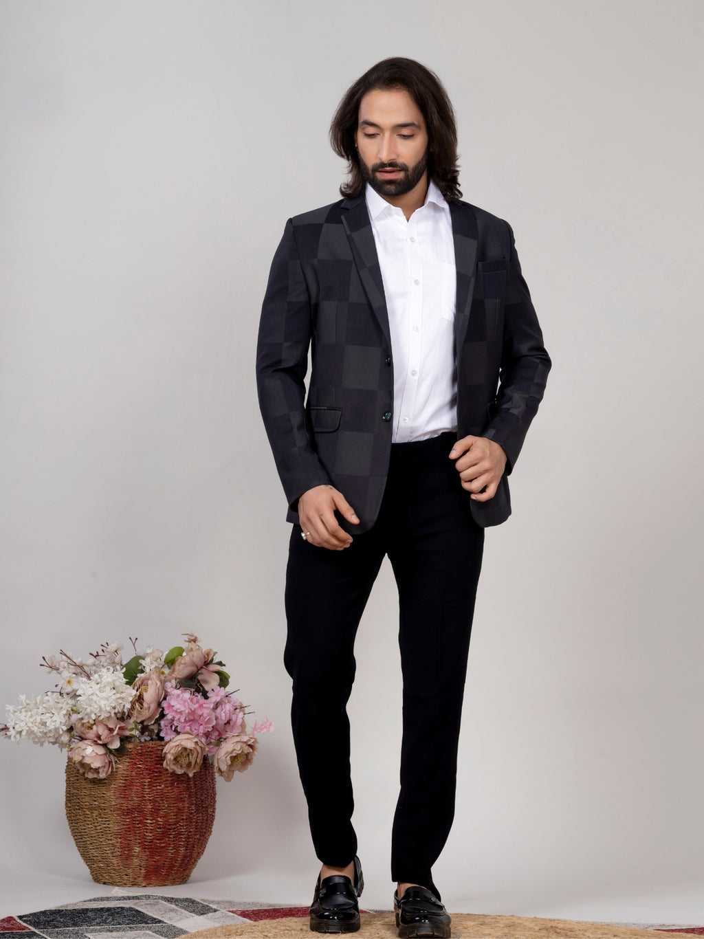 Man wearing a black suit with a white shirt, standing against a plain background with a vase of flowers.