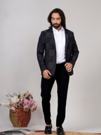 Man wearing a black suit with a white shirt, standing against a plain background with a vase of flowers.