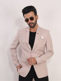 A man wearing a light pink blazer over a black shirt and black pants, accessorized with round sunglasses and a white pocket square, posing against a plain white background.