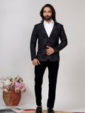 A man modeling a black checkered blazer with a white shirt and black trousers, standing against a plain light background next to a decorative basket of flowers.