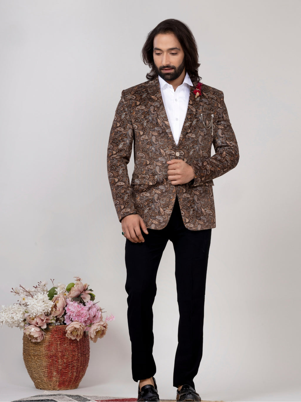 A man wearing a brown and beige patterned blazer over a white shirt and black trousers, standing in front of a light gray studio background with a basket of flowers at his side.