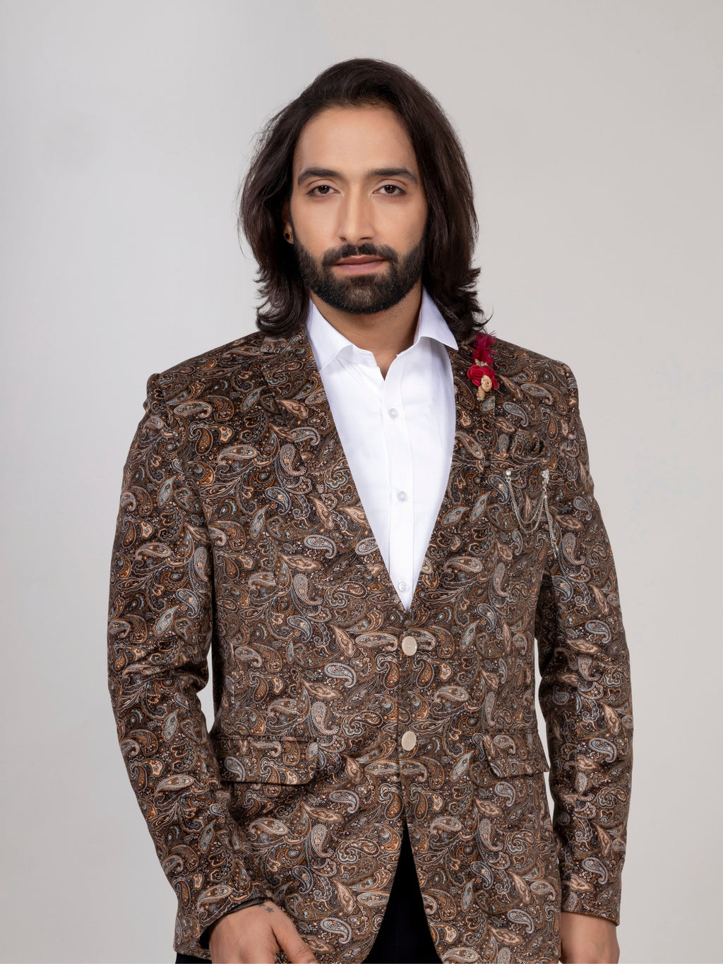 A man with long dark hair and a beard wearing a brown paisley patterned blazer over a white shirt, standing against a light gray studio background.