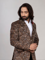 A man with long dark hair and a beard wearing a brown paisley patterned blazer over a white shirt, standing against a light gray studio background.