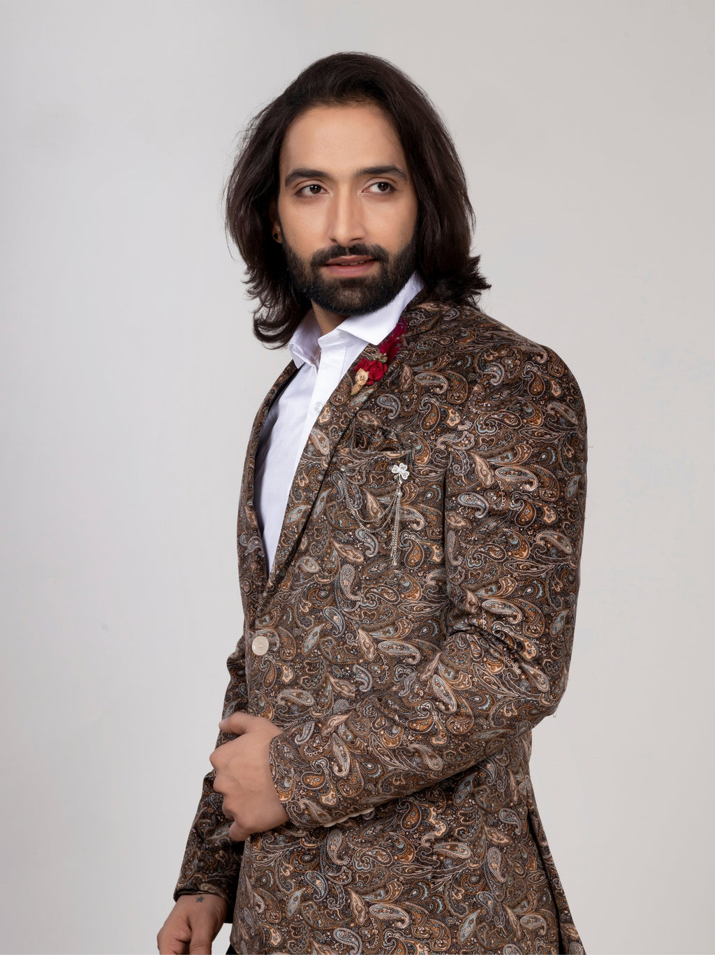 A man with long dark hair and a beard wearing a brown paisley patterned blazer over a white shirt, standing against a light gray studio background.