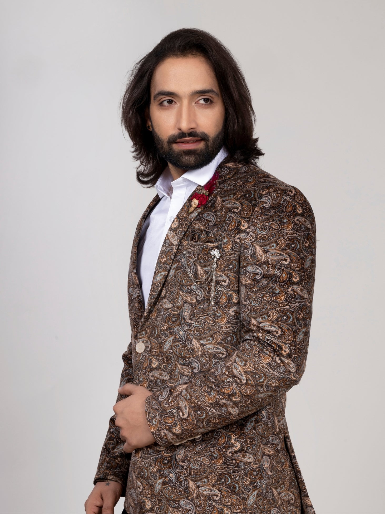 A man with long dark hair and a beard wearing a brown paisley patterned blazer over a white shirt, standing against a light gray studio background.