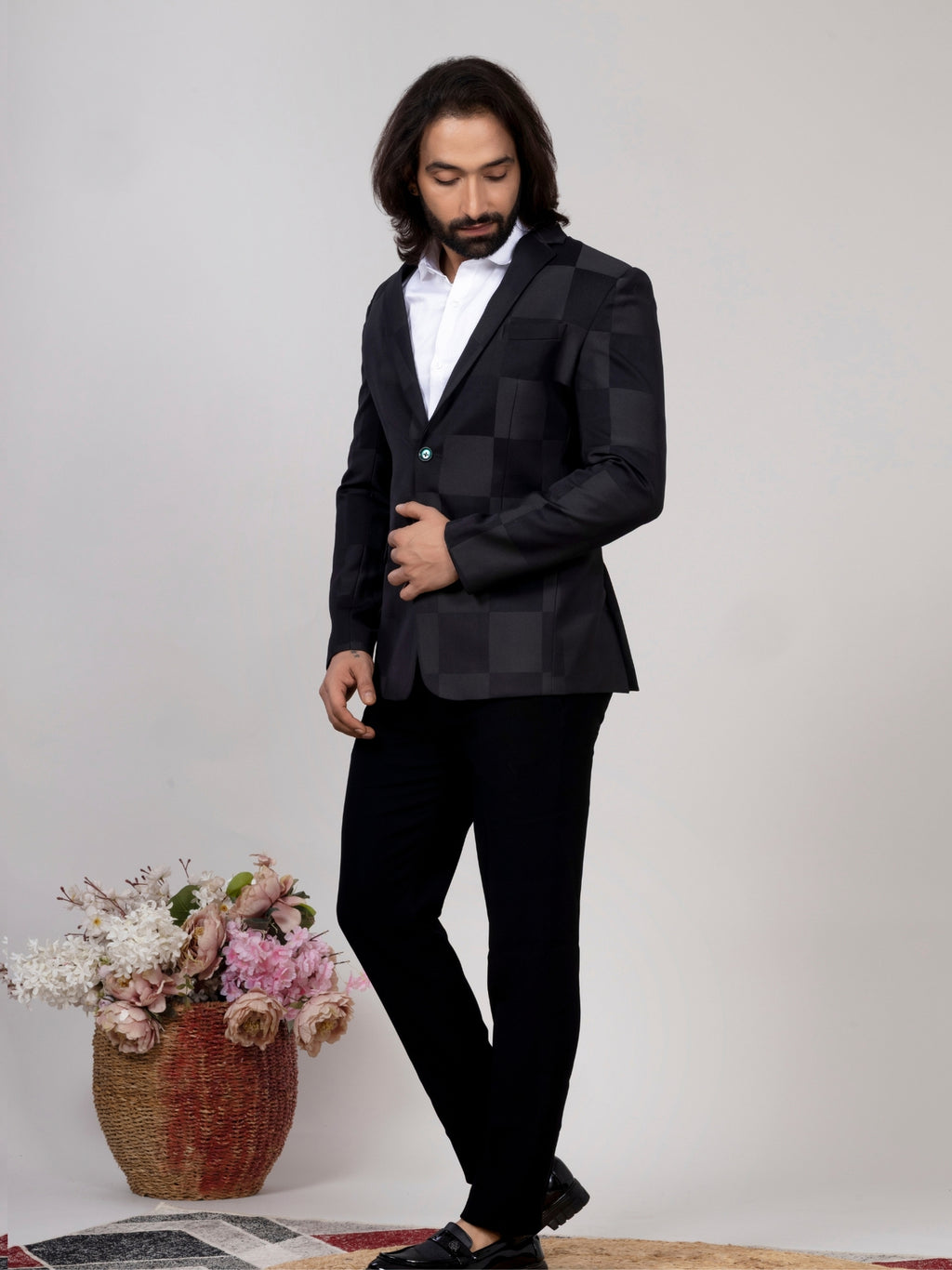 A man wearing a black checkered blazer, white shirt, and black trousers, standing in a studio setting next to a decorative basket of flowers.