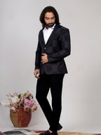 A man wearing a black checkered blazer, white shirt, and black trousers, standing in a studio setting next to a decorative basket of flowers.