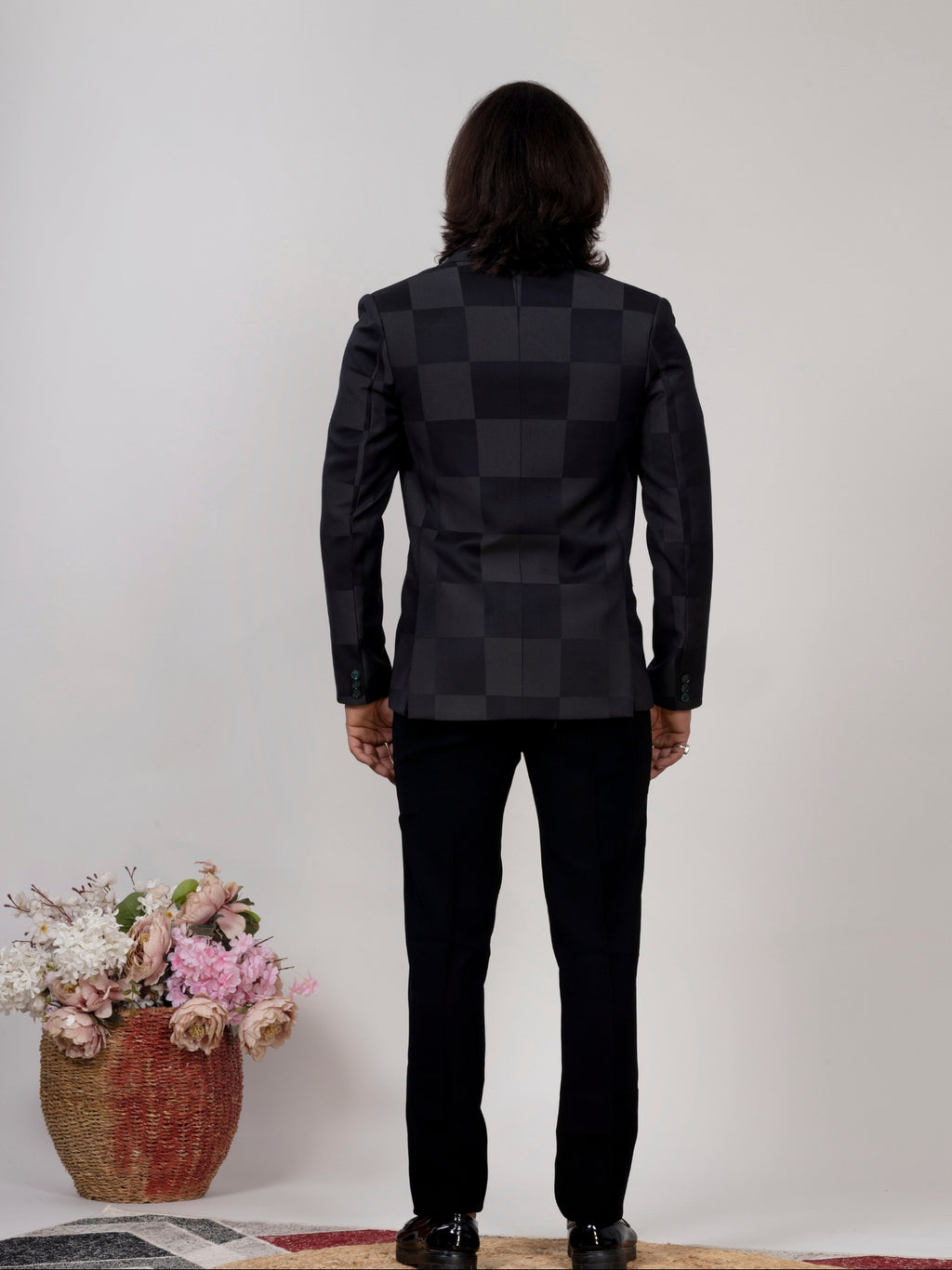 A man viewed from behind wearing a black checkered blazer and black trousers, standing against a plain light gray wall with a basket of flowers on the floor to the left.