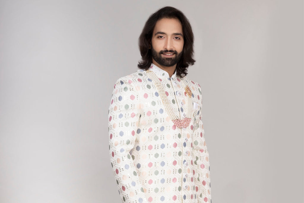 Man wearing a patterned kurta with white pants standing next to a vase of flowers on a checkered floor.