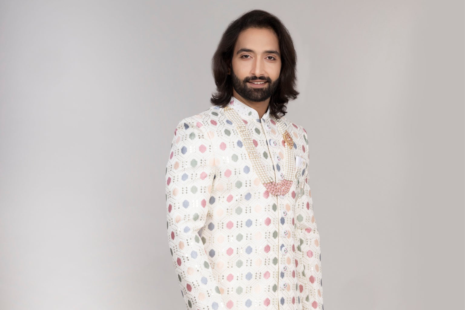Man wearing a patterned kurta with white pants standing next to a vase of flowers on a checkered floor.