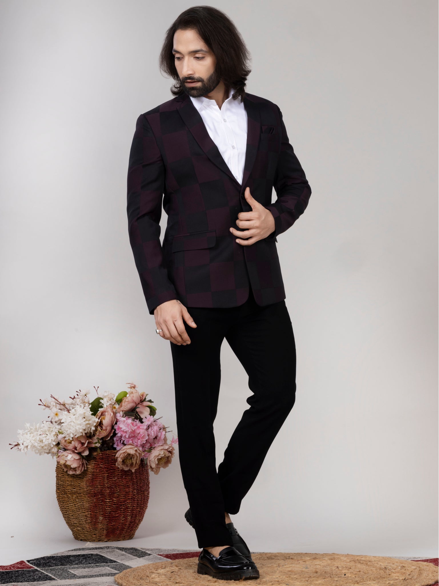 Man wearing a dark suit with a white shirt, standing against a plain background with a vase of flowers.