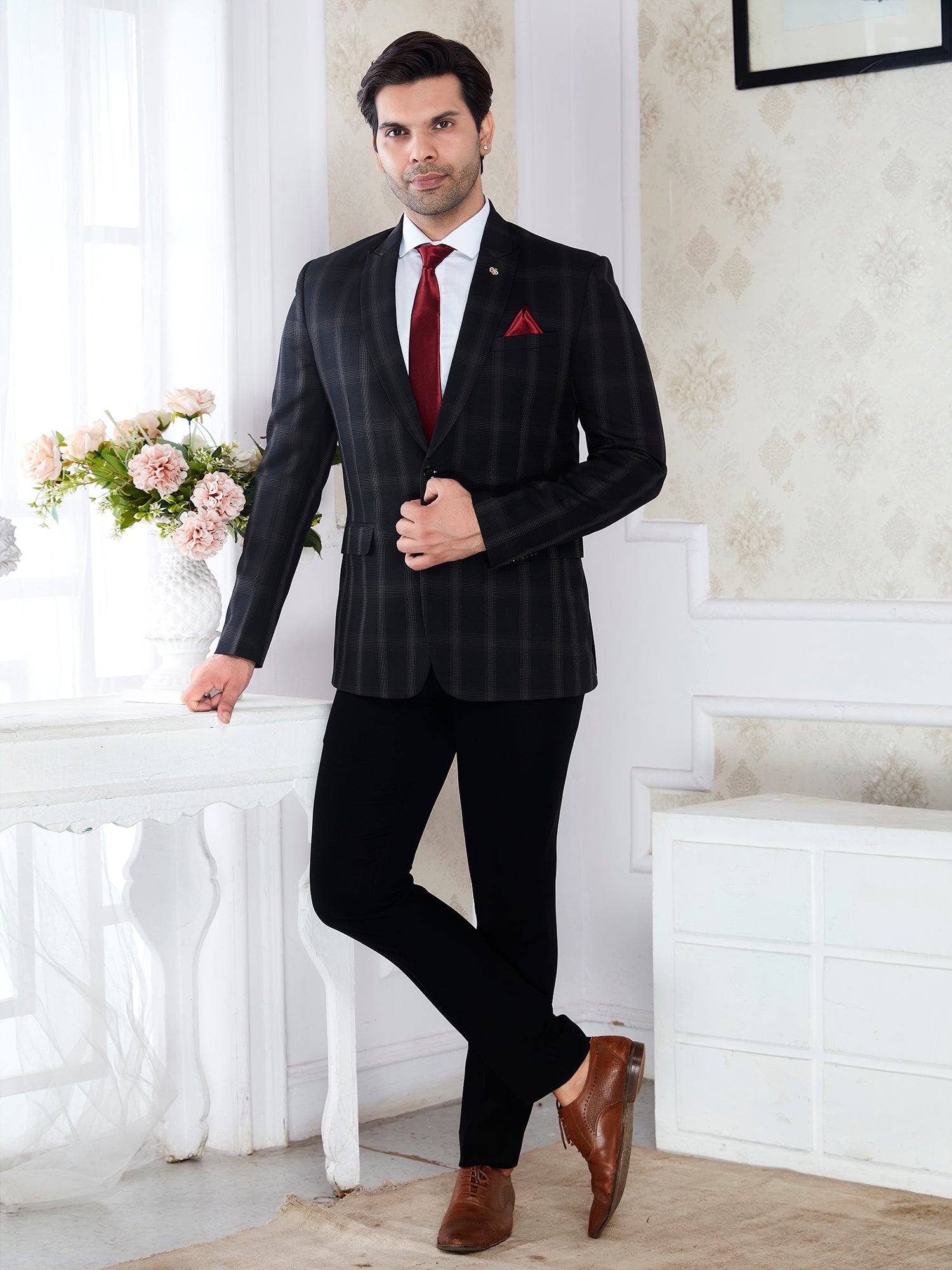 Man in a black pinstripe suit with a red tie standing in a room with white walls and a table with flowers.