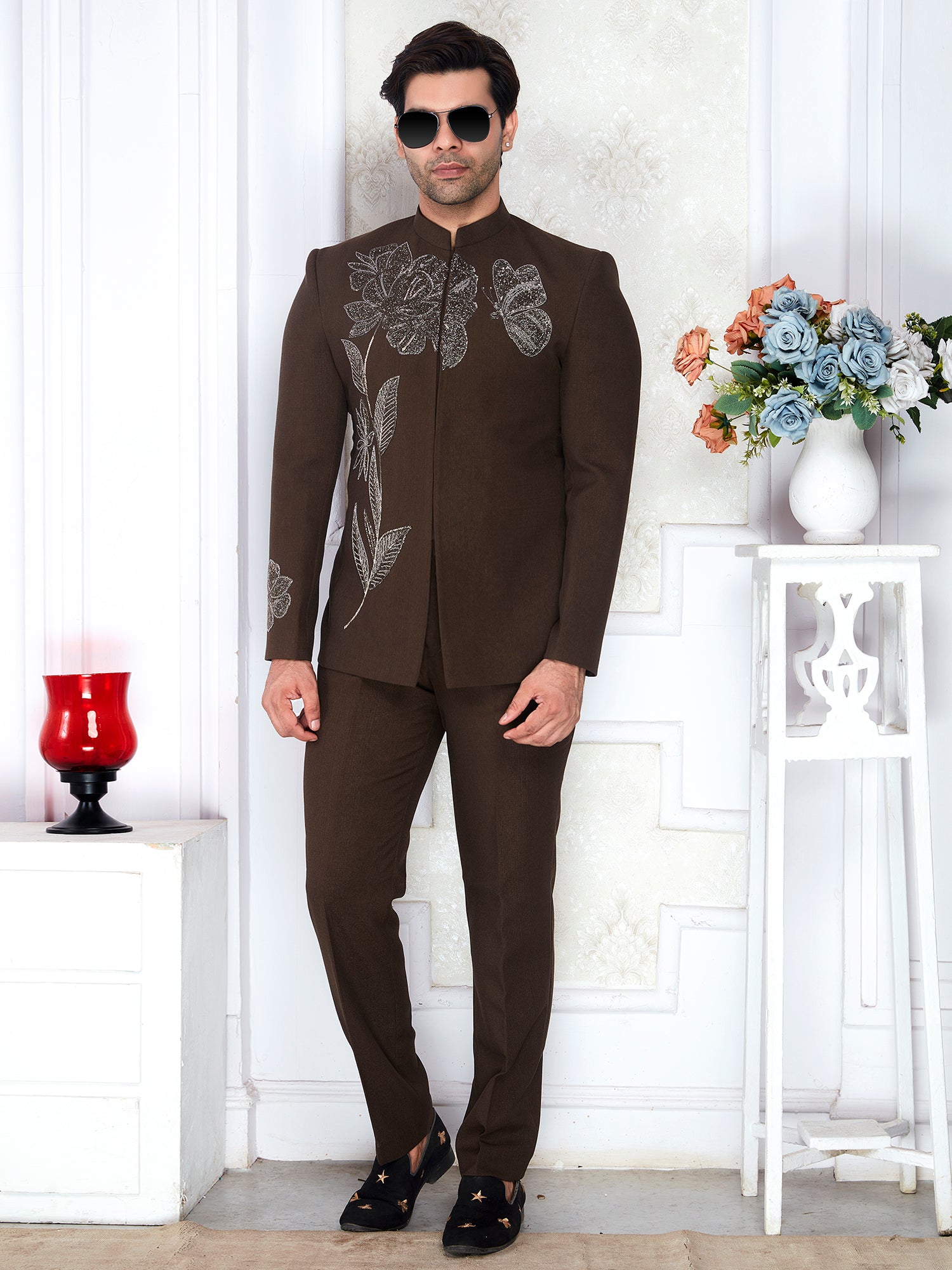 Man wearing a brown embroidered suit standing in a room with decorative elements.