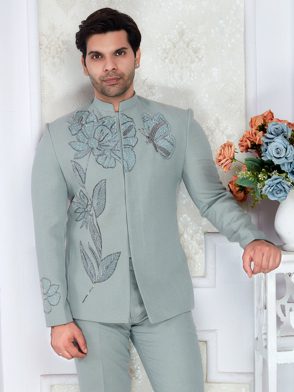 Man wearing a light gray embroidered suit standing against a white wall with decorative elements.