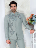 Man wearing a light gray embroidered suit standing against a white wall with decorative elements.