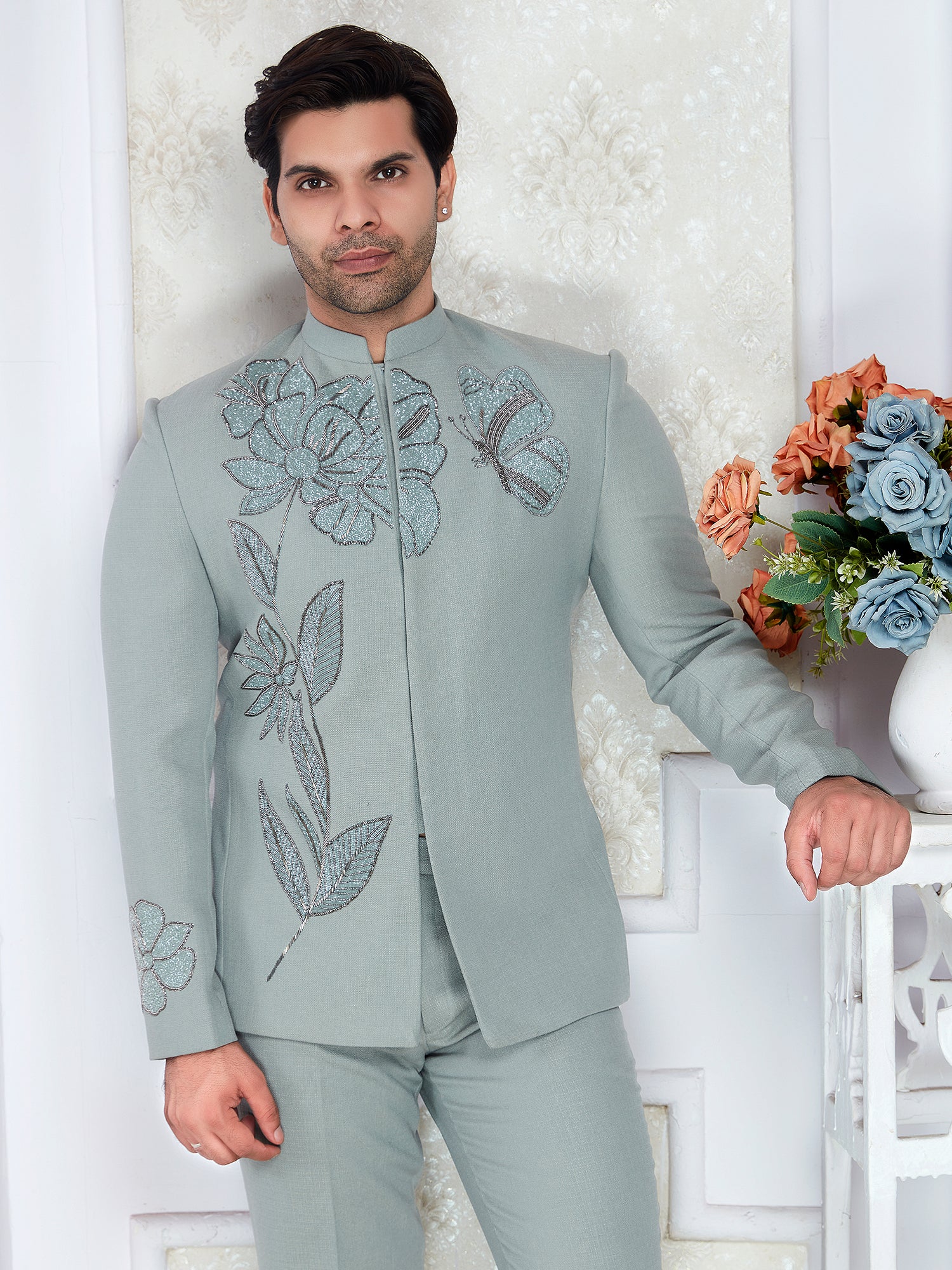 Man wearing a light gray embroidered suit standing against a white wall with decorative elements.