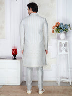 Man wearing a light gray sherwani in a room with white walls and a small table with flowers.