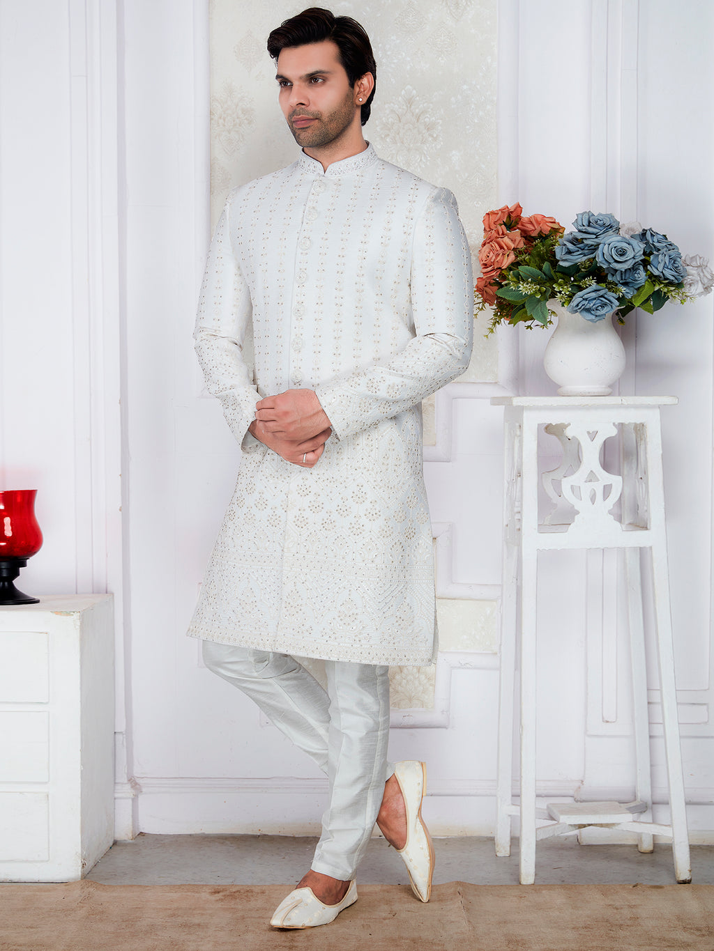 Man wearing a white sherwani standing in a room with a decorative vase and flowers.