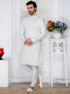 Man wearing a white sherwani standing in a room with a decorative vase and flowers.