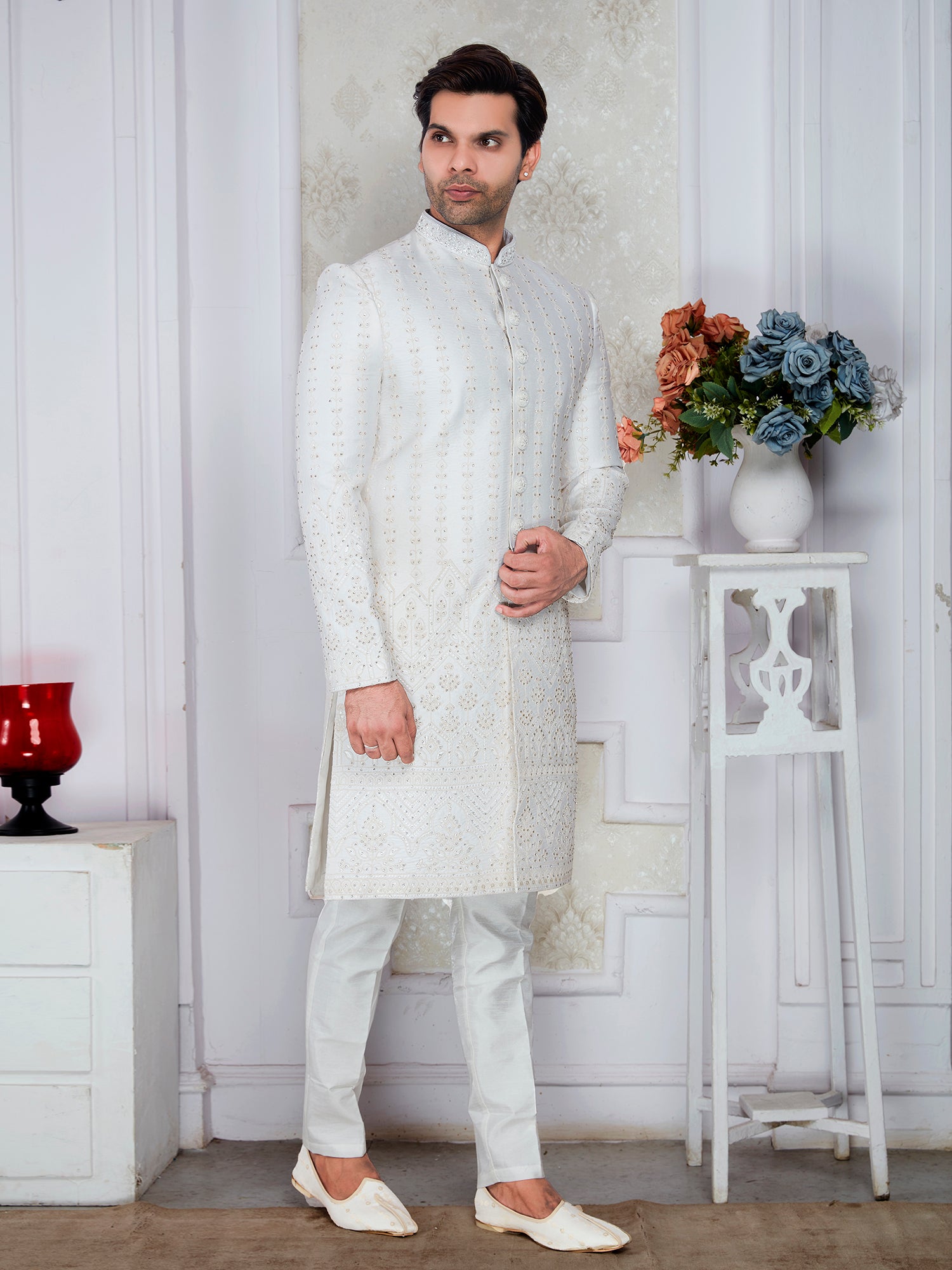 Man wearing a white sherwani in a decorated room with flowers and decor items.