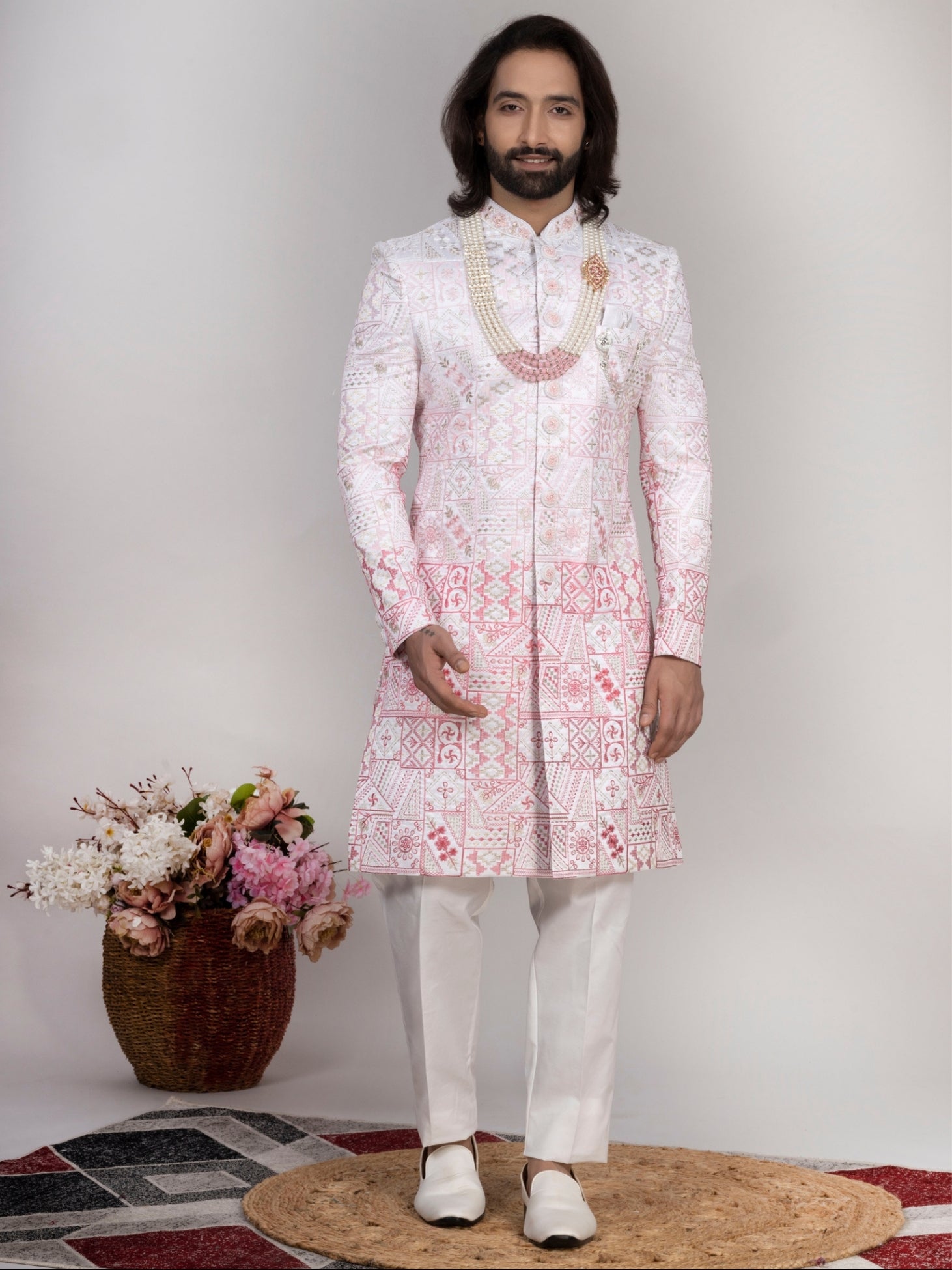 A man modeling a traditional Indian sherwani in light pink and white with intricate embroidery, paired with white trousers and white shoes, standing in front of a neutral studio background.