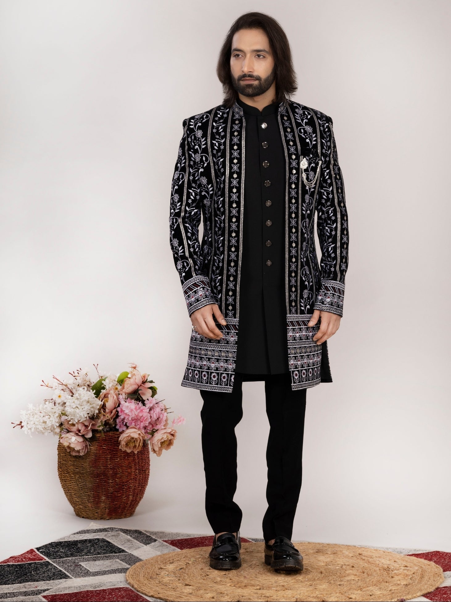 A man modeling a black long sherwani jacket with intricate white and gray geometric patterns, paired with a black kurta, black trousers, and black shoes, standing against a plain off-white background.