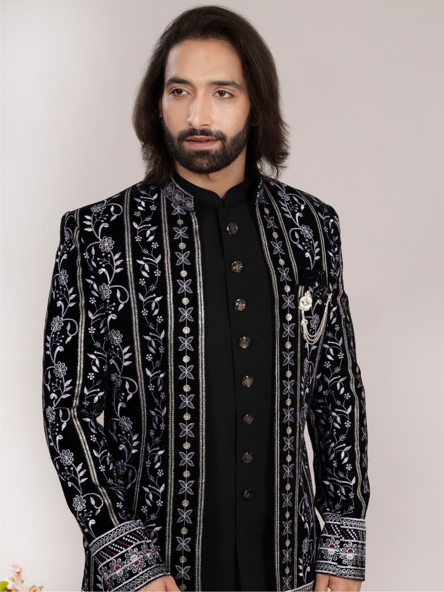 A man wearing a black velvet sherwani jacket with intricate silver and white floral embroidery, paired with a black kurta, against a plain light gray background.