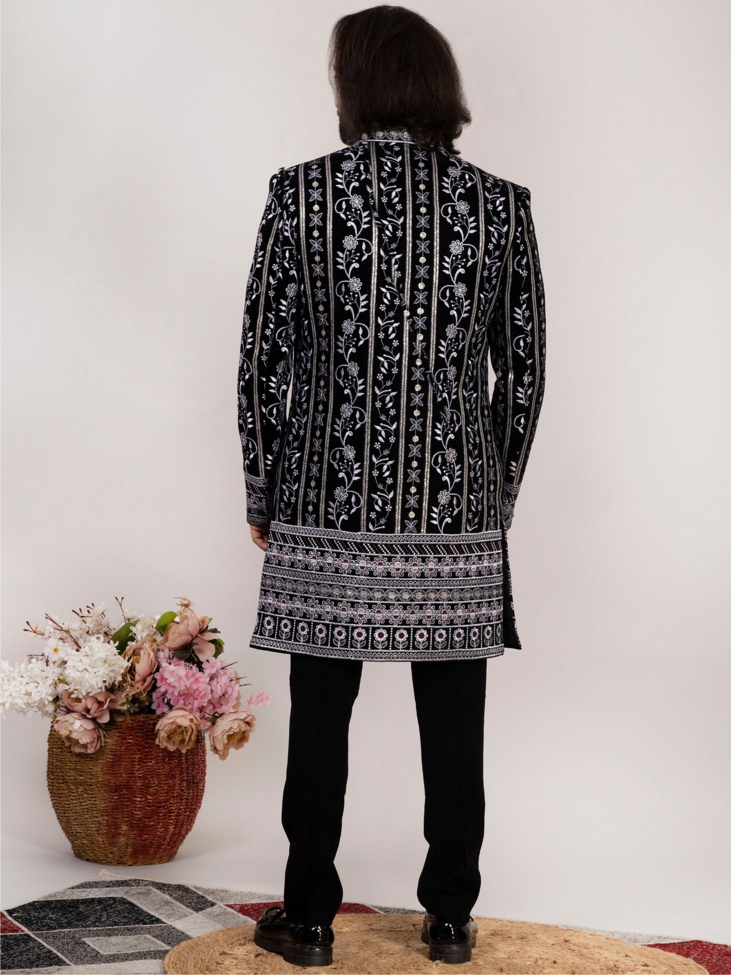 A person shown from the back wearing a black long kurta or tunic with intricate white floral and geometric embroidery, paired with black trousers and black shoes. The setting is indoors against a plain light wall, with a woven basket of flowers on the left and a round rug underfoot.